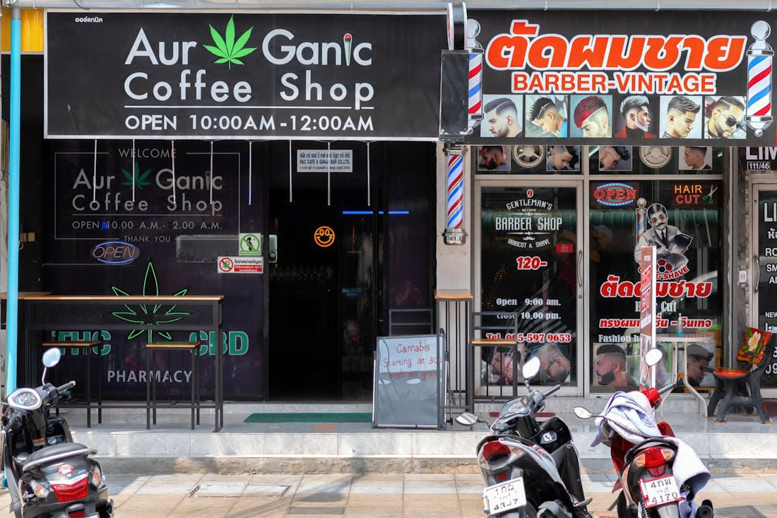 Cannabis-Shop in Thailand mit leuchtendem Schild ‚Aur Ganic Coffee Shop‘ neben einem Barber-Shop, Motorräder davor geparkt.