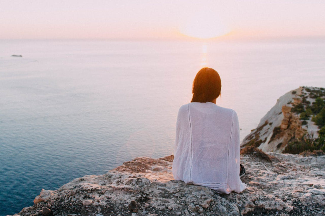 Person sitzt ruhig auf Felsen und blickt auf das Meer bei Sonnenuntergang – Symbol für Achtsamkeit, Ruhe und inneres Gleichgewicht