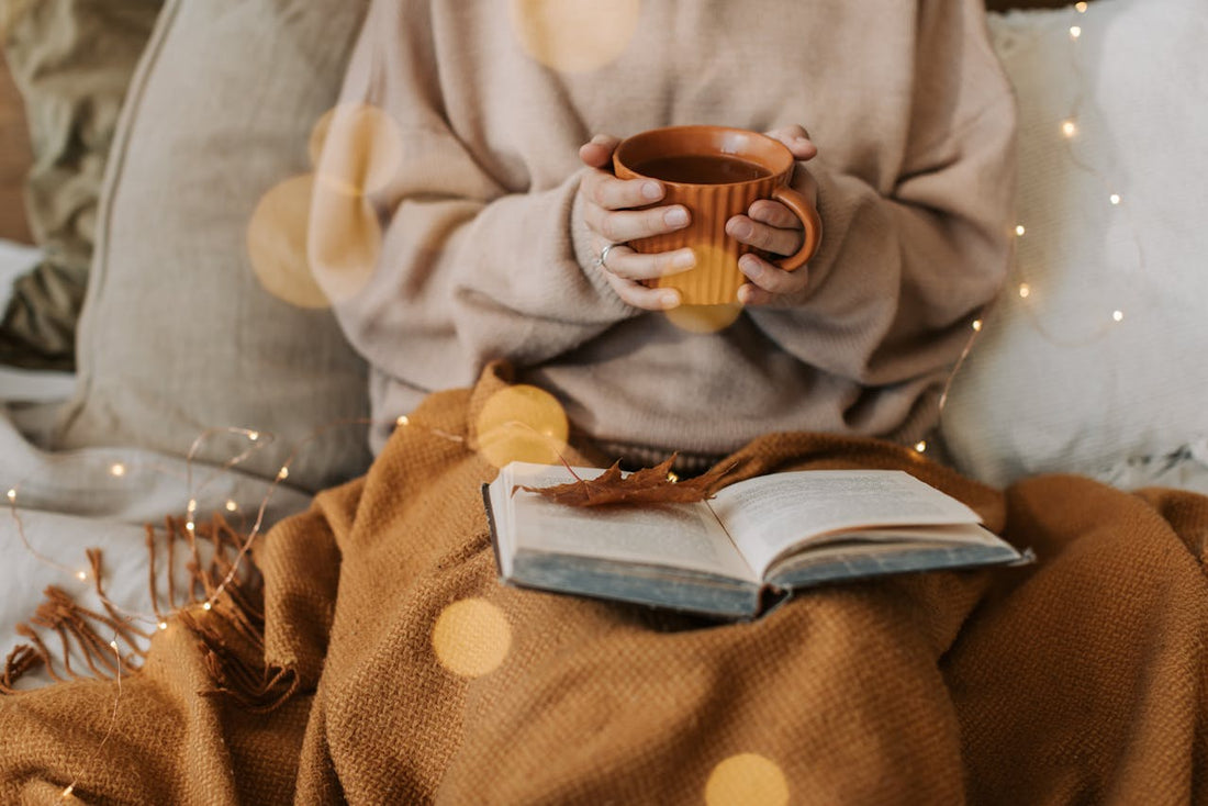 Person mit warmer Tasse Tee, Decke und offenem Buch – stimmungsvolle Abendroutine als Sinnbild für entspannteren Schlaf